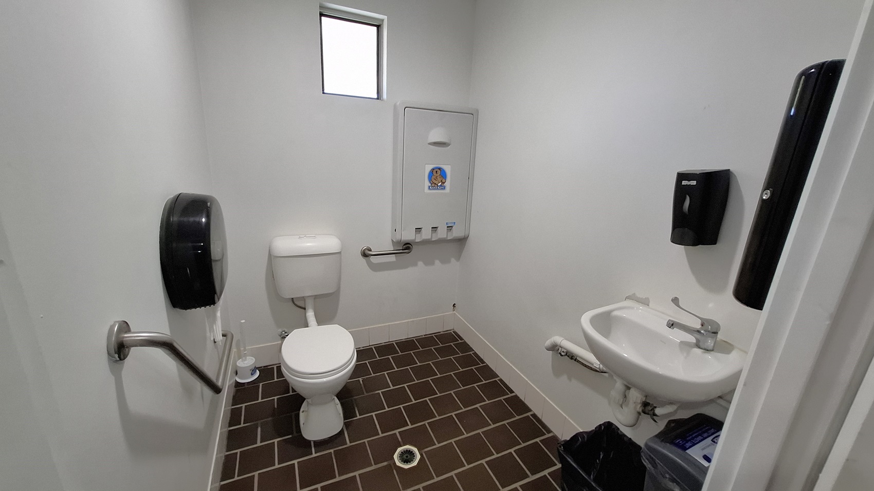 On the left, a white toilet is positioned next to a wall-mounted paper towel dispenser. A grab bar is installed beside the toilet for support. In the centre, the floor is covered with dark brown tiles arranged in a grid pattern, leading to a drain in the middle. To the right, a small sink is mounted on the wall, with a tap and a waste bin underneath. A small window above the toilet allows light into the space. The walls are painted white, and the ceiling is also white.