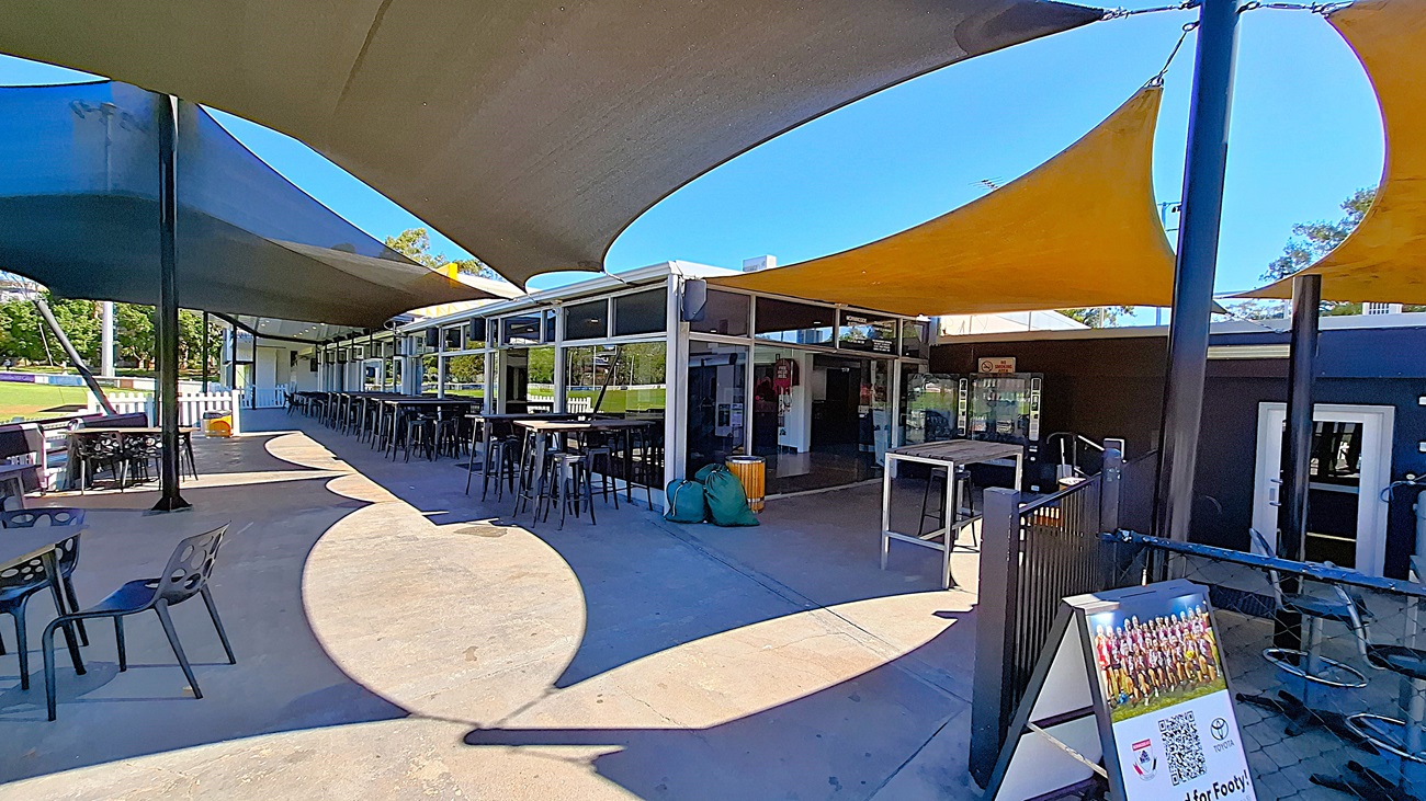 On the left, a wooden bin is positioned next to a green bag, while a glass door leads into the club. To the right, two vending machines are visible, displaying various snacks and drinks. In the centre, the entrance opens to a spacious area with wooden flooring, leading to tall tables and chairs arranged throughout. The ceiling is covered with a tan awning, providing shade. Above the entrance, a sign displays the club's name and trading hours.