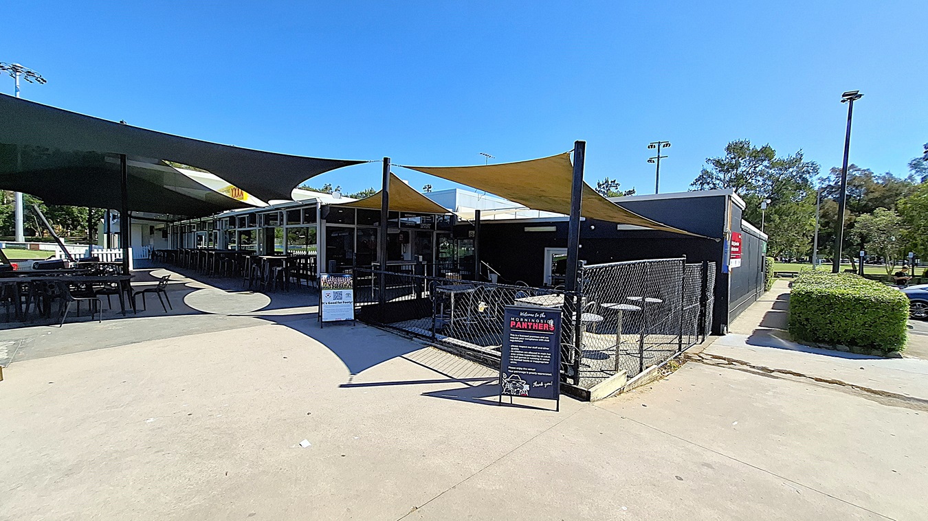 On the left, a series of black tables and chairs are arranged under a large green shade sail, providing outdoor seating. The floor is made of smooth concrete, leading towards a fenced area. In the centre, the entrance to the building features large glass windows, allowing visibility into the interior. To the right, a sign stands on the ground, displaying information about the venue. The ceiling is formed by the shade sails, which are yellow and black,
