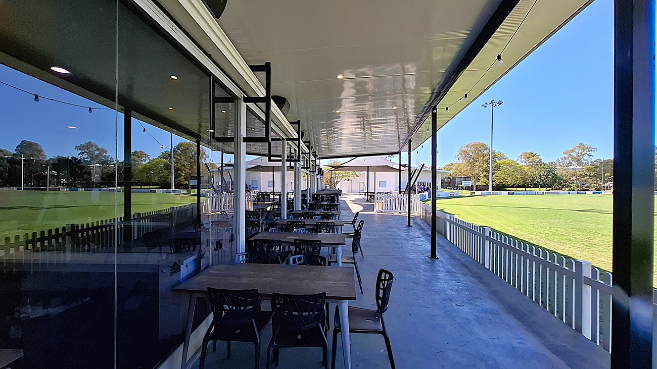 On the left, a row of wooden tables with black chairs is positioned under a covered area, with large glass windows reflecting the surroundings. The floor is smooth and grey, extending towards the right. In the background, a green field is visible, bordered by a white picket fence. To the right, additional seating is arranged, leading to a building with white walls. The ceiling is flat and white, with overhead lights providing illumination.