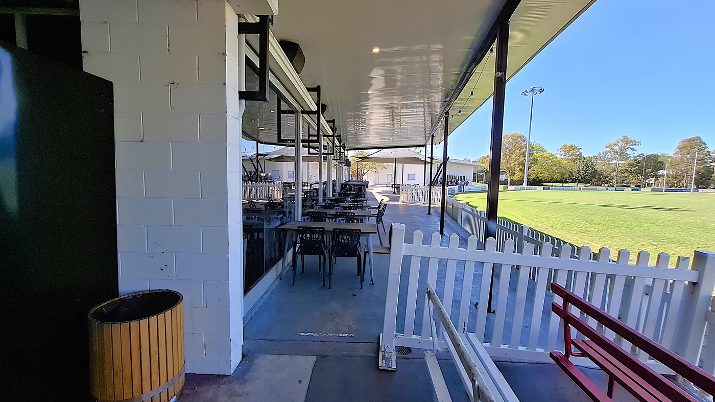 On the left, a green wall is visible next to a wooden bin, while a series of tables and chairs are arranged along the covered area. The floor is smooth and grey, extending towards the right. In the background, a white picket fence borders a green field, with several buildings visible beyond it. To the right, a red bench is positioned against the fence. The ceiling is flat and white, with overhead lights.