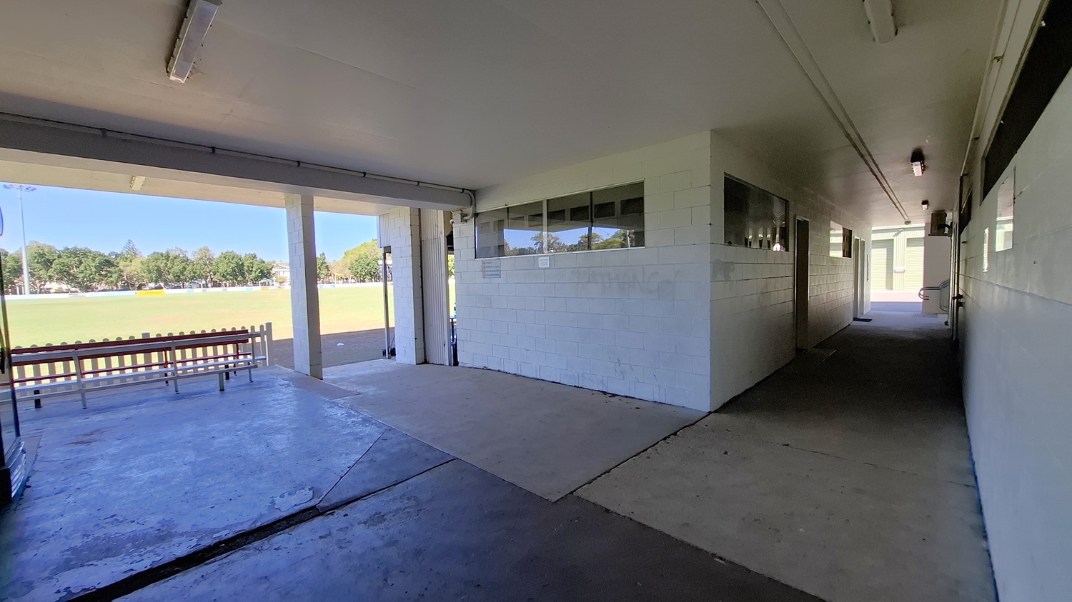 On the left, a concrete wall features a series of windows, allowing light into the corridor. A bench with a red seat is positioned against the wall, facing an open area. The floor is a mix of concrete and tiles, leading towards a grassy field visible in the background. To the right, a narrow corridor extends, with doors along the wall and a light fixture above. The ceiling is flat and white, with fluorescent lights providing illumination throughout the space.