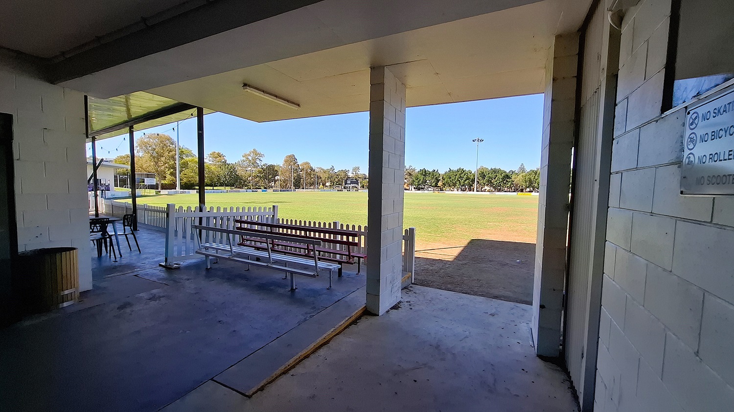 On the left, a wooden bin is positioned next to a wall, with a small table and chairs visible nearby. The floor is a combination of concrete and tiles, leading to an open area. In the foreground, a white picket fence separates the space from a grassy field, which extends into the background. To the right, a wall features a sign with various rules. The ceiling is flat and white, with overhead lights.