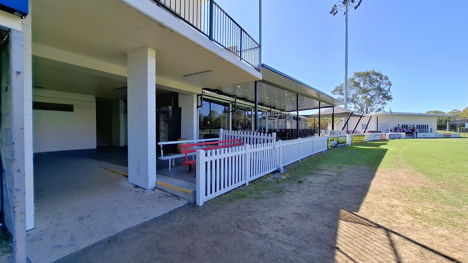 On the left, a concrete area leads to a covered space with a white railing and a red bench. The floor is a mix of concrete and grass, transitioning to a grassy field in the foreground. In the background, a large glass structure features multiple windows, providing views of the interior seating. To the right, another building is visible, with a similar design. The ceiling above the covered area is flat and extends outwards, providing shelter while allowing light to enter through the glass panels.