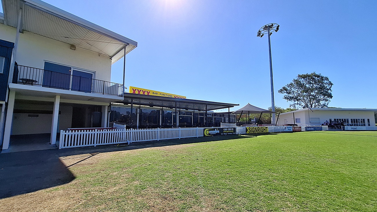 On the left, a two-storey building features a balcony with black railings and large windows. Below, a covered area with glass panels provides seating. The floor is a mix of grass and concrete, leading to a well-maintained green field in the foreground. To the right, another building with white walls and large windows is visible, along with a tall light pole. The ceiling of the covered area is flat and extends outwards. A white picket fence runs along the front.