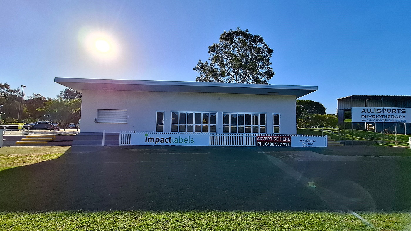 On the left, a modern building features large glass windows and a white sign with "impact labels" displayed prominently. Steps lead up to the entrance, which is flanked by a grassy area. The floor is a mix of grass and concrete, extending towards the right. To the right, another building is visible with a sign for "All Sports Physiotherapy." The ceiling of the main building is flat and white, providing a clean, functional appearance.