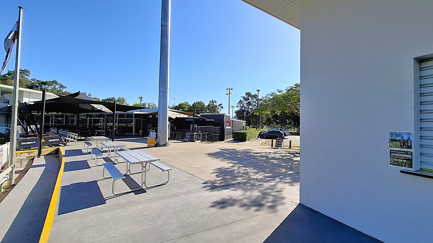 On the left, a series of picnic tables are arranged on a concrete surface, with a yellow edge marking the area. A tall light pole stands nearby, and a shaded seating area is visible in the background, featuring large glass windows. The floor is smooth concrete, extending towards a grassy area in the foreground. To the right, a white wall with a small window displays a poster. The ceiling above the seating area is flat and extends outwards.