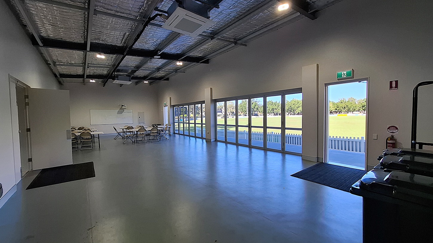 On the left, a small meeting area features several folding chairs and tables arranged neatly. A whiteboard is mounted on the wall, and a door leads to another room. The floor is smooth and grey, extending towards the right side. To the right, large glass doors open to a grassy area outside, with a white picket fence visible beyond. The ceiling is high with exposed beams and insulation, fitted with overhead lights. An exit sign is mounted above the door, indicating the way out.