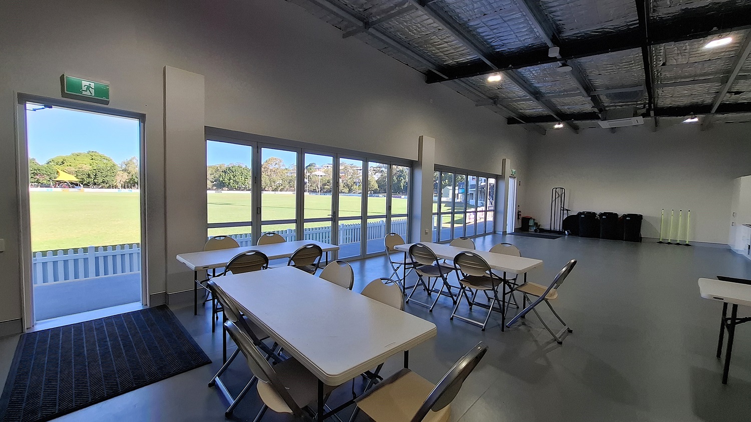 On the left, a doorway leads outside, framed by a grey wall. A black mat is positioned at the entrance, and large glass doors provide a view of a grassy field beyond. Several white tables and folding chairs are arranged in the middle of the room, with a smooth grey floor beneath. To the right, a wall features a small window and a few bins stacked against it. The ceiling is high with exposed beams and insulation, fitted with overhead lights .