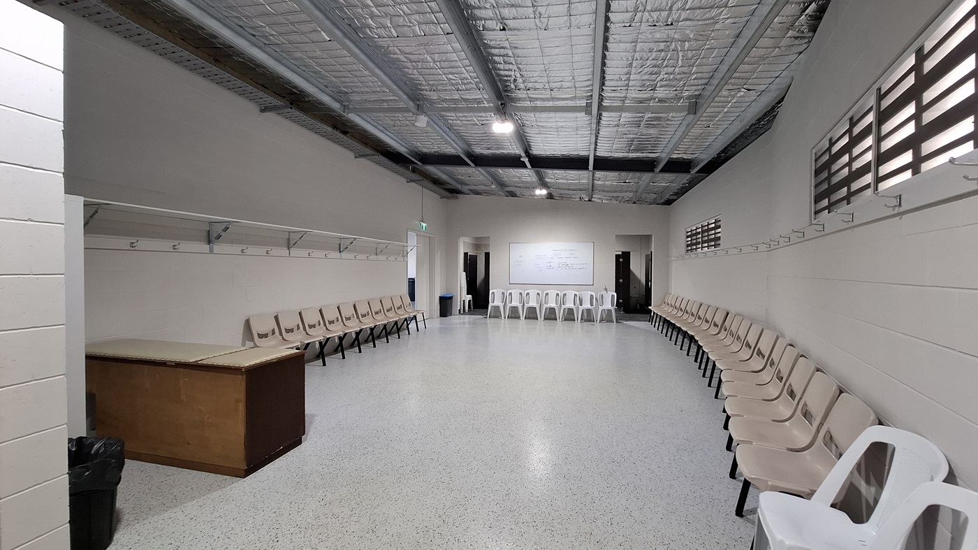 On the left, a wooden table is positioned against the wall, with a black bin nearby. Above, a row of hooks is mounted for hanging items. The floor is smooth and light-coloured, extending throughout the room. In the middle, a whiteboard is mounted on the wall, with several chairs arranged in a semi-circle around the space. To the right, more chairs are lined up against the wall, and a doorway leads to another area. The ceiling is flat with exposed beams and insulation, fitted with overhead lights