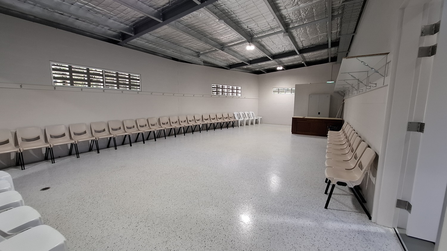 On the left, a row of beige chairs is arranged along the wall, with a few white chairs interspersed. Above, small windows allow light to enter, and a rail runs along the wall for hanging items. The floor is smooth and polished, reflecting the light. In the middle, the space is open, leading to a wooden counter against the right wall. The ceiling is high with exposed beams and insulation, fitted with overhead lights.