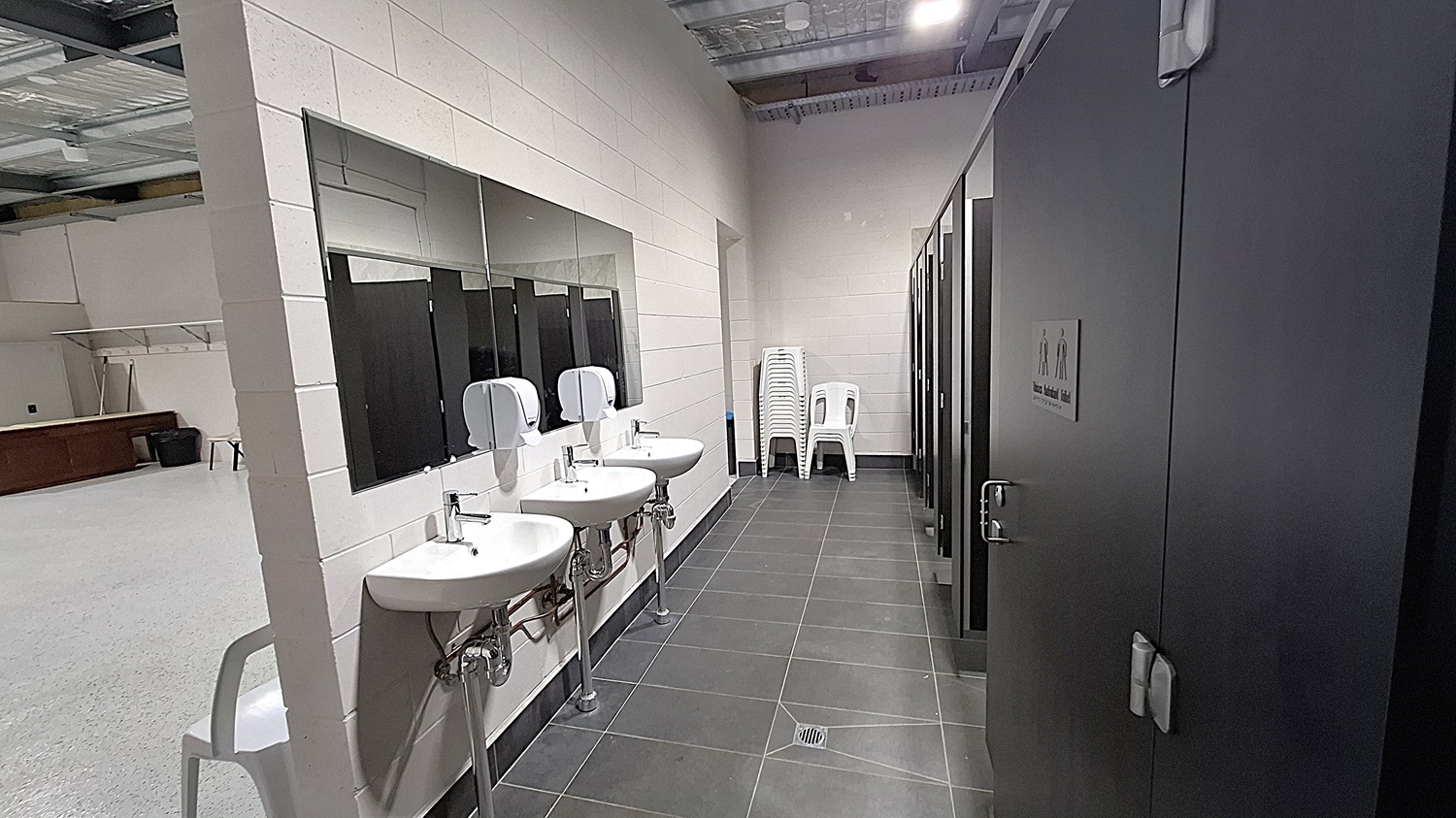 On the left, three white sinks are mounted on the wall, each with a soap dispenser above. A large mirror spans the length of the sinks. The floor is dark grey tiles, extending towards the right. To the right, several black cubicle doors are visible, each with a sign indicating male and female facilities. A stack of white plastic chairs is positioned against the wall. The ceiling is high with exposed metal beams and panels, fitted with overhead lights.