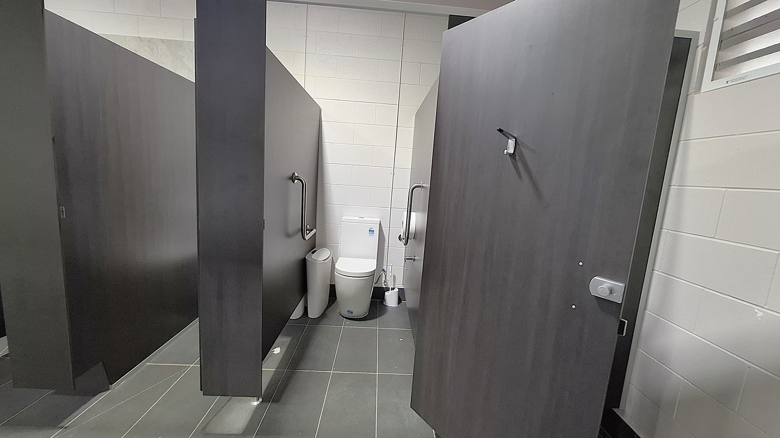 On the left, a dark wooden cubicle door stands closed, with a grab rail mounted beside it. Adjacent to it, a white toilet is positioned against the wall, with a small bin nearby. The floor is dark grey tiles, extending throughout the space. To the right, another cubicle door is slightly ajar, revealing a similar setup. The ceiling is flat and white, with ventilation grilles visible, providing a clean and functional environment.