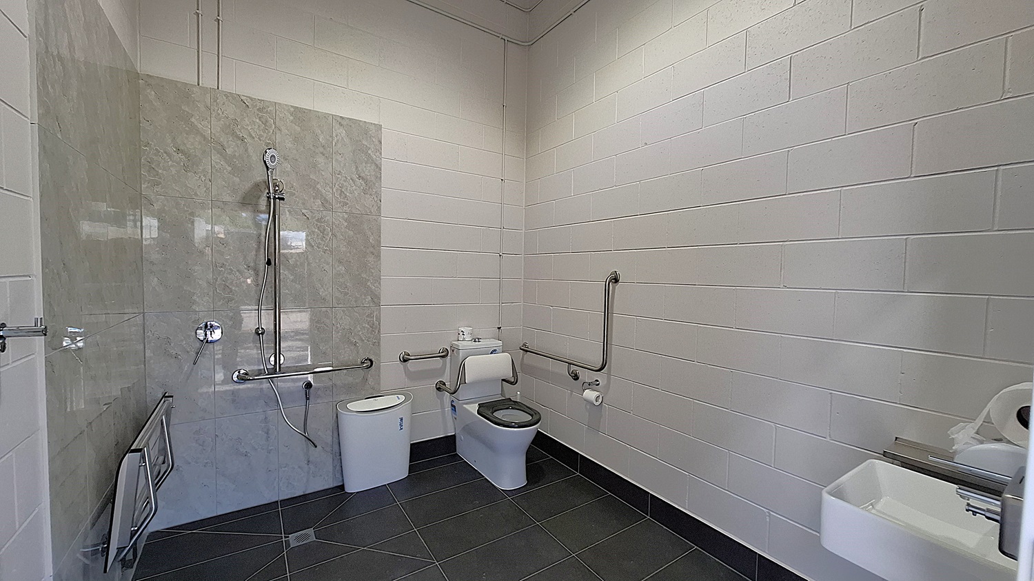 On the left, a shower area features a wall of grey tiles with a handheld showerhead mounted above. A grab rail is installed beside the shower. Adjacent to it, a white toilet is positioned with a roll of toilet paper mounted on the wall. The floor is dark grey tiles, extending throughout the space. To the right, a small sink is mounted on the wall, with a paper towel dispenser nearby. The ceiling is flat and white, with exposed pipes running along it.