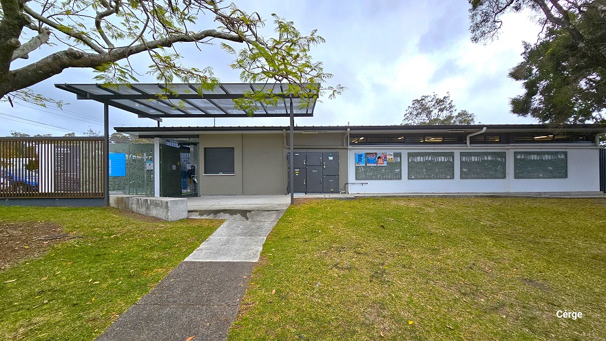 Grey and white cemented pathway leading to the facility entrance. There is a large open space in the front of the facility that is covered with grass. A grey metal locker cabinet is placed against the right wall, which is sun-protected by a skylight roofing supported by a metal roof frame. A few meters away are four windows with some small adverts in the first window frame.