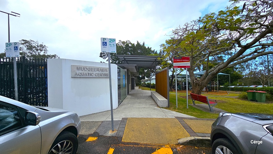 Parking for people with disability is marked by a white parking sign with the letter P, accessibility logo, and the word Only printed on the flat sign board. Beside is the 2-hour parking sign with green print including date details. On the left side is the name of the facility, detailed on a white wall, while on the right is a brown fence with skylit roofing that covers the cemented smooth pathway at the center. At the back of the red bench is a tall red signboard.