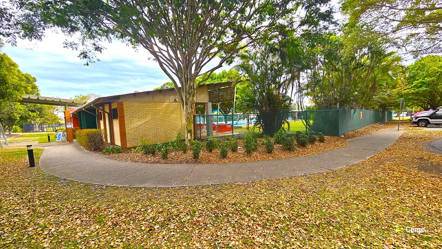 A concrete pathway with a rough surface connects the car park to a low-set cream brick building with light brown brick foundation edges. Black safety bollards can be seen on the side of the concrete pathway. Covering the landscape is dried leaf litter. Small green plants with parallel leaf venation can be seen on the right, just before the building.