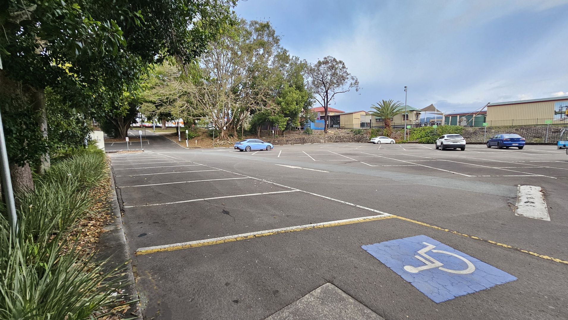 A nearly empty car park with a few cars scattered around. In the foreground, a marked disabled parking space is visible. On the left, there are dense trees and bushes lining the edge of the car park. In the background, a row of buildings and a fence are visible, with some trees and a palm tree nearby. The surface of the car park is asphalt, with white lines marking the parking spaces.