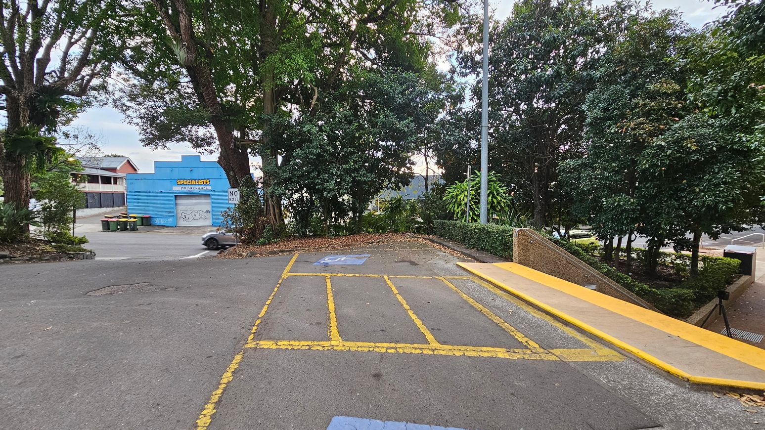 A parking area with yellow lines and a blue disabled parking symbol on the ground. On the right, there's a yellow ramp leading downwards, bordered by bushes and trees. In the background, a blue building with a sign reading "SPECIALISTS" is visible, along with several bins. A car is partially visible near the building. Tall trees surround the area, providing shade and greenery.