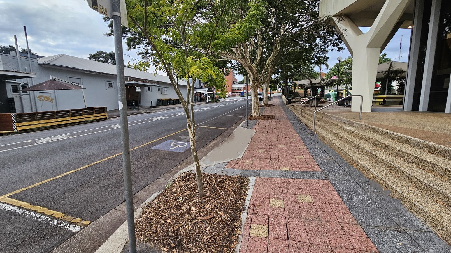 On the left, a road with a bicycle lane marked by a symbol, bordered by a pavement with trees planted in square soil patches. A building with a grey roof and a small outdoor seating area is visible. On the right, a wide pavement with red and grey tiles leads to a building with large steps and a modern architectural design. Trees line the pavement, and a metal railing runs alongside the steps. In the background, more buildings and a bus stop are visible.