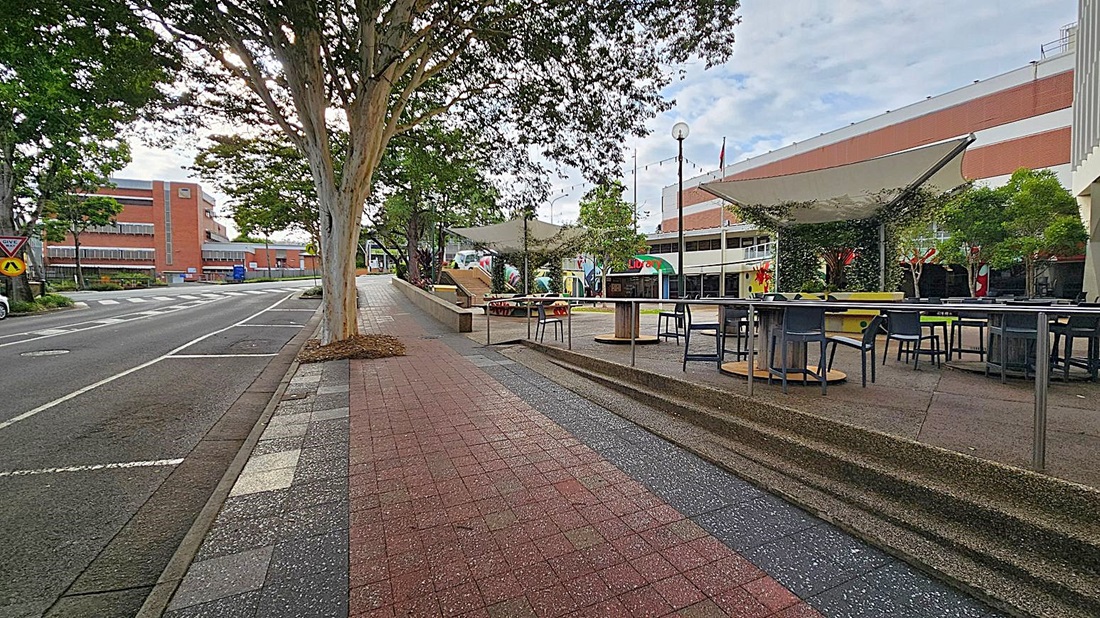 A street scene with a footpath on the left featuring a metal railing and a grey chair. Trees line the path, providing shade. On the right, a road with parked cars and a row of buildings, including a small structure with a red awning. Several bins are lined up against a building. The pavement is a mix of red and grey tiles, and a large tree trunk is visible in the foreground.