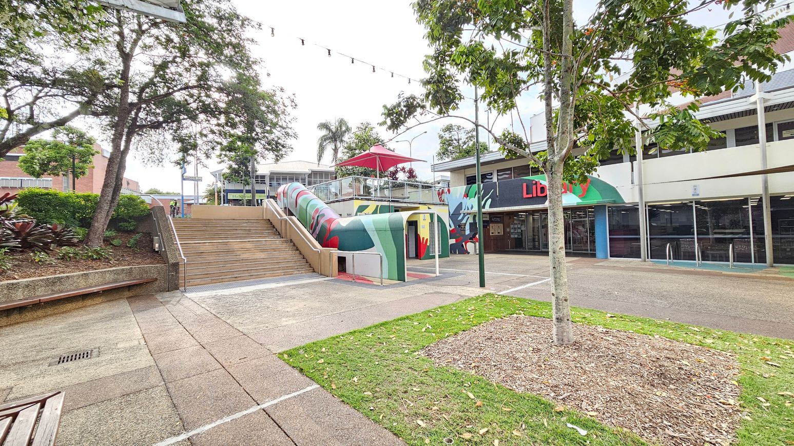 A public space with a library on the right, featuring a green and red awning. In the centre, a colourful, curved structure with a red umbrella on top leads to a staircase. On the left, there are trees and shrubs with a bench along a raised garden bed. The ground is paved with concrete, and a small patch of grass with a tree is in the foreground. Overhead, string lights are visible.