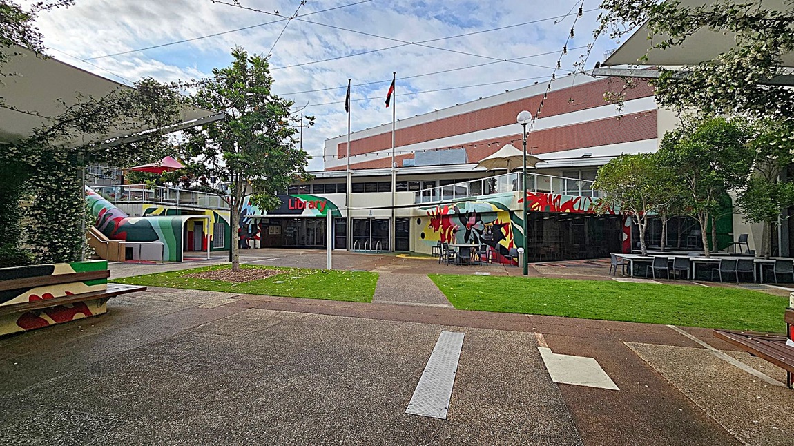A courtyard with a library in the background. The library has colourful murals on its walls and a sign reading "Library" above the entrance. Two flagpoles stand in front of the building. On the left, a covered area with greenery and a bench. On the right, tables and chairs under trees. The ground is a mix of grass and paved paths, and string lights are strung overhead.