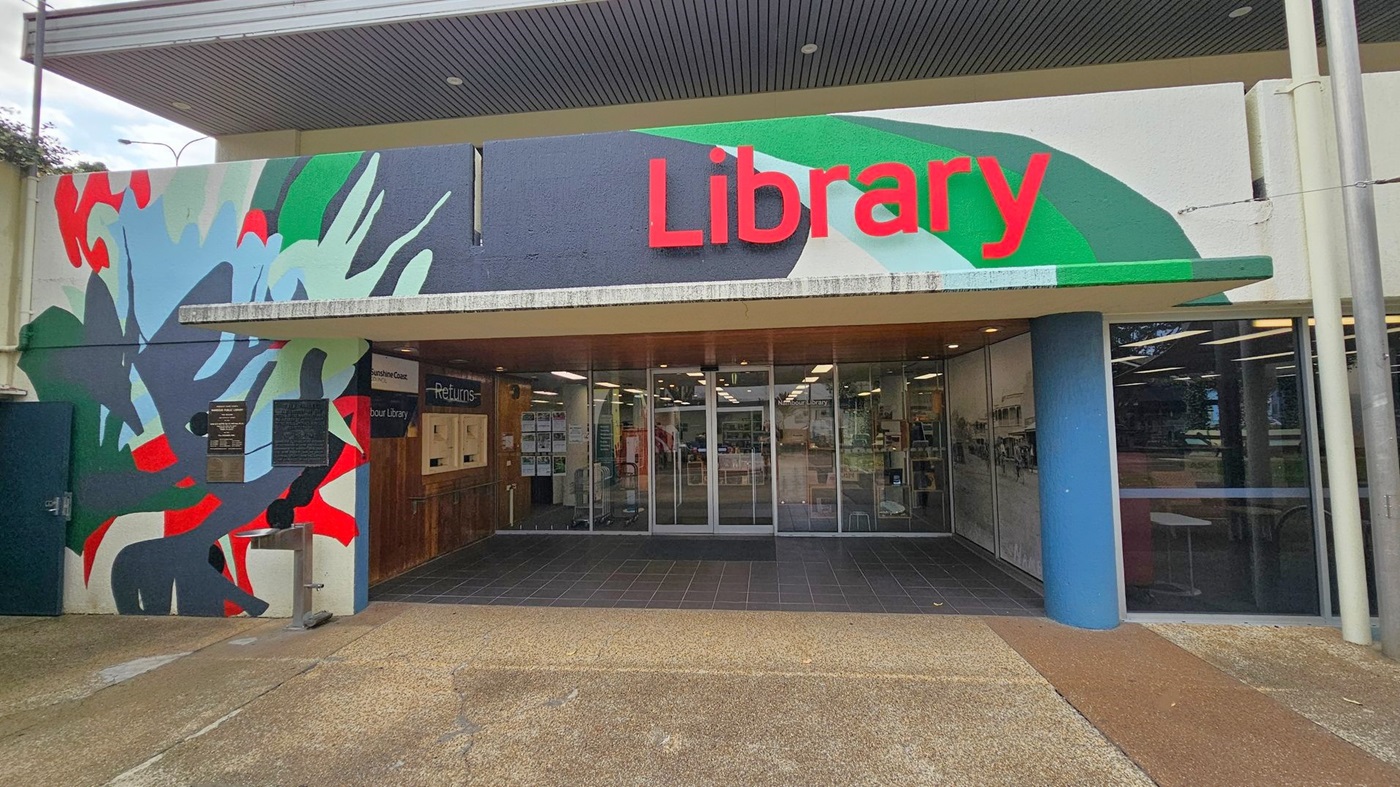 Entrance to a library with a colourful mural on the left featuring abstract shapes in red, green, and blue. The word "Library" is prominently displayed in red on the right. Glass doors lead into the building, with a returns section visible on the left. The floor outside is a mix of concrete and tiles, and the ceiling is dark with recessed lighting.
