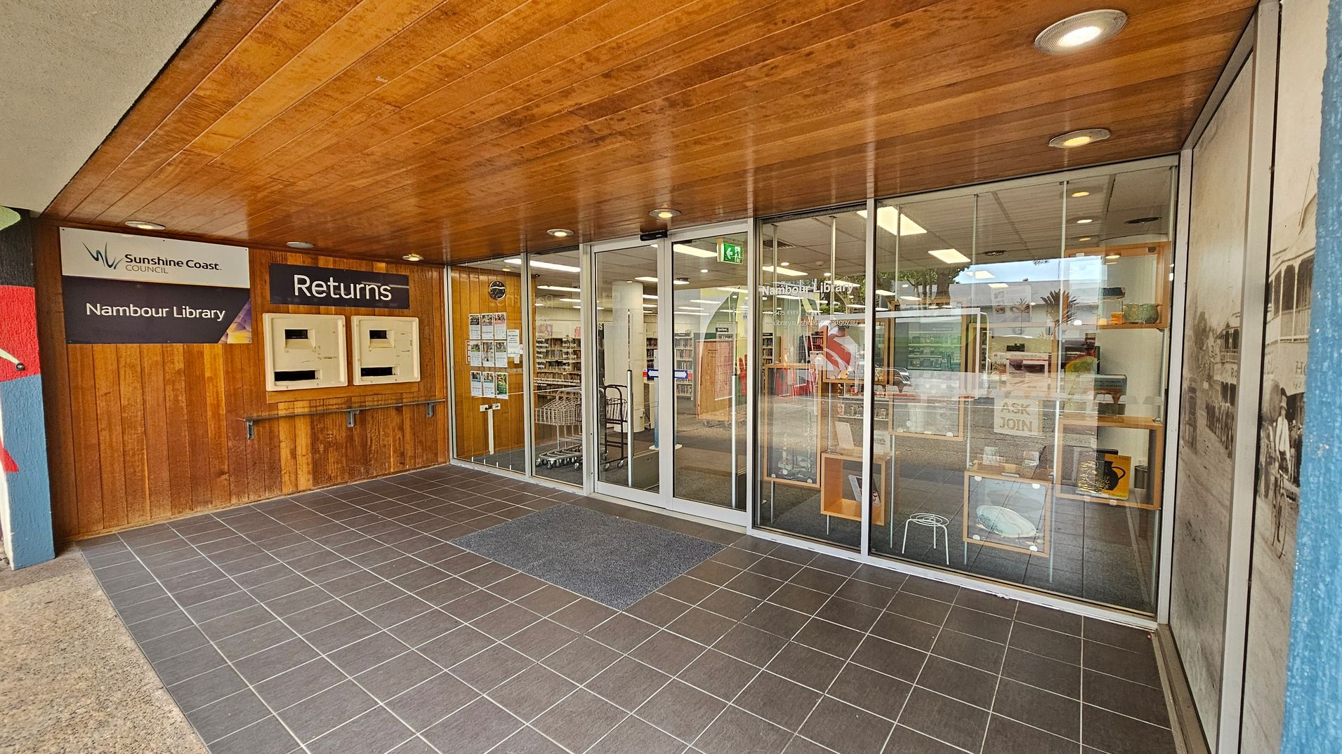 Entrance to Nambour Library with a wooden ceiling and tiled floor. On the left, there are two return slots under a "Returns" sign, and a "Sunshine Coast Council Nambour Library" sign. The glass doors in the centre lead into the library, revealing bookshelves and a cart inside. Posters and notices are displayed on the glass. On the right, a large window shows a historical photo and some library displays.