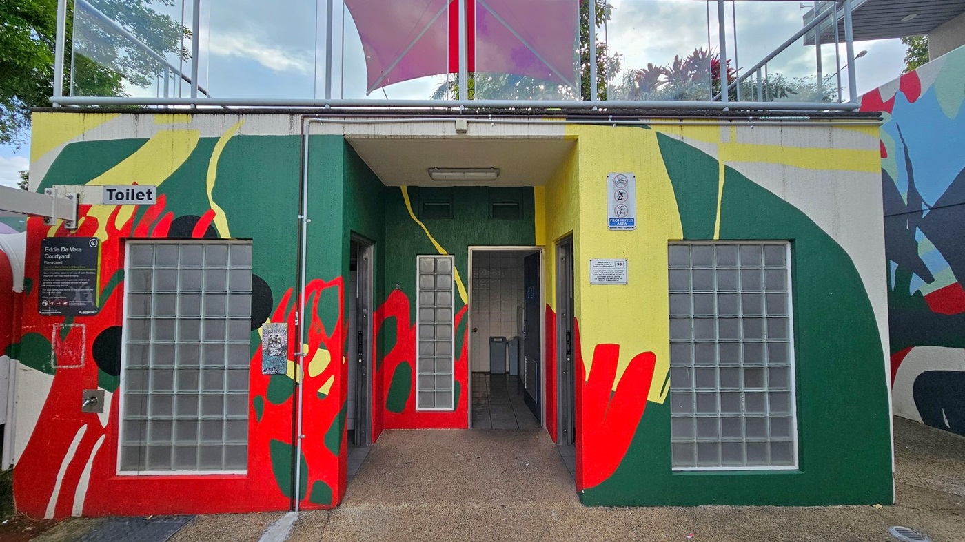A public toilet building with a colourful mural featuring green, red, yellow, and black abstract shapes. The entrance is in the centre, flanked by glass block windows. A sign above the left side reads "Toilet," and there's a plaque for "Eddie De Vere Courtyard" below it. The ground is paved, and a pink canopy is visible above. Trees and a railing are in the background.
