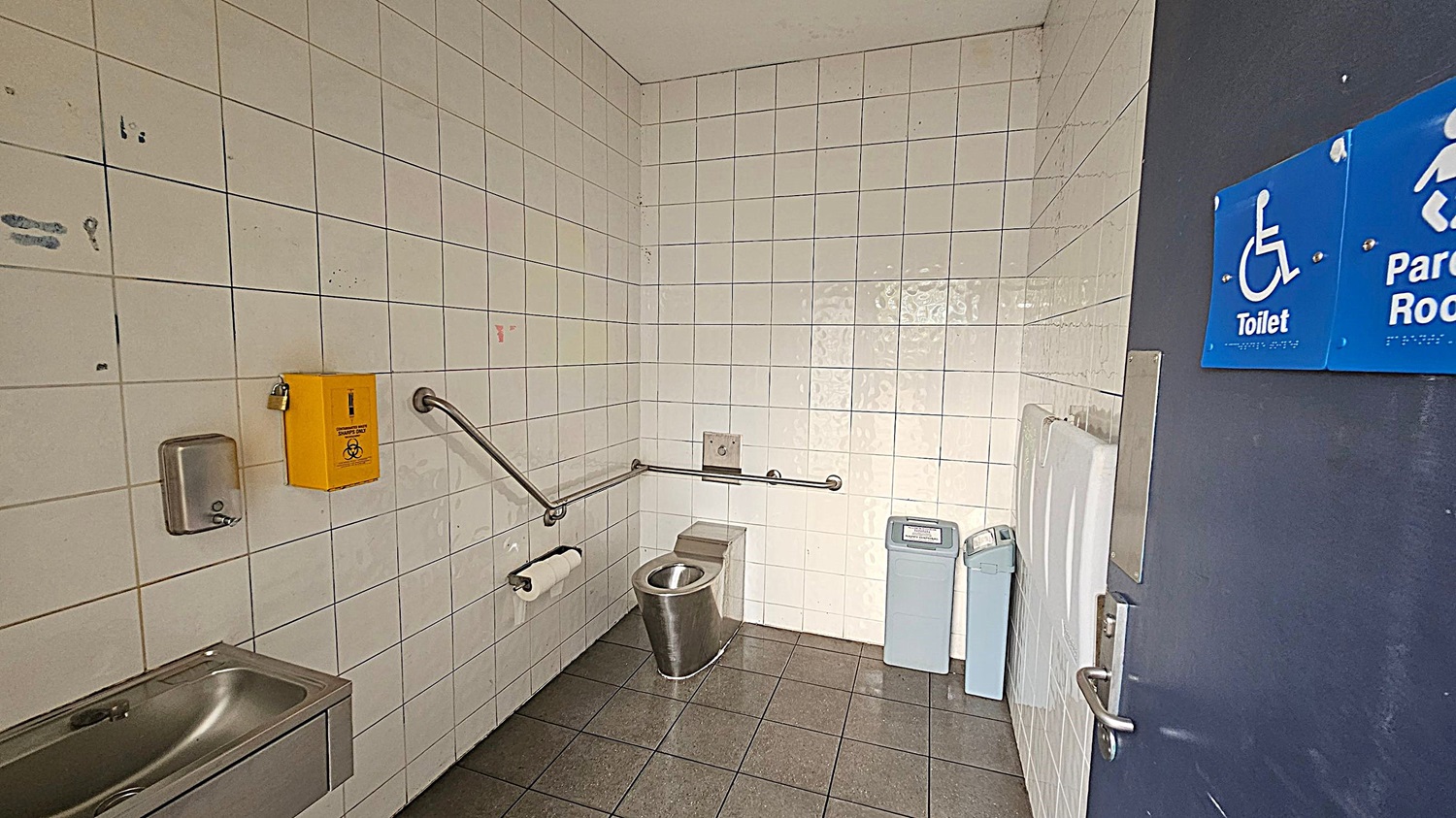 A public accessible toilet with white tiled walls and a grey tiled floor. On the left, there's a stainless steel sink with a soap dispenser above it and a yellow biohazard disposal box. A metal grab rail runs along the wall, with a toilet roll holder attached. In the centre, a stainless steel toilet is positioned against the wall. On the right, there are two bins and a baby changing station. The door has a blue sign indicating it's a toilet and parent room.