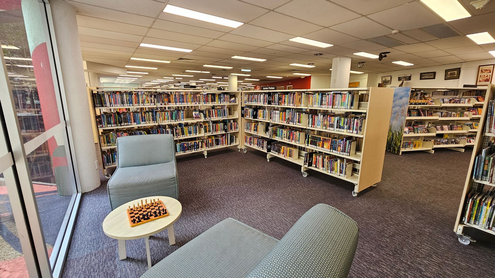 A library interior with bookshelves filled with books on the right and in the background. In the foreground, there are two grey upholstered chairs and a small round table with a chessboard on it. The floor is carpeted in a dark grey colour. Large windows on the left allow natural light to enter. The ceiling is lined with fluorescent lights.