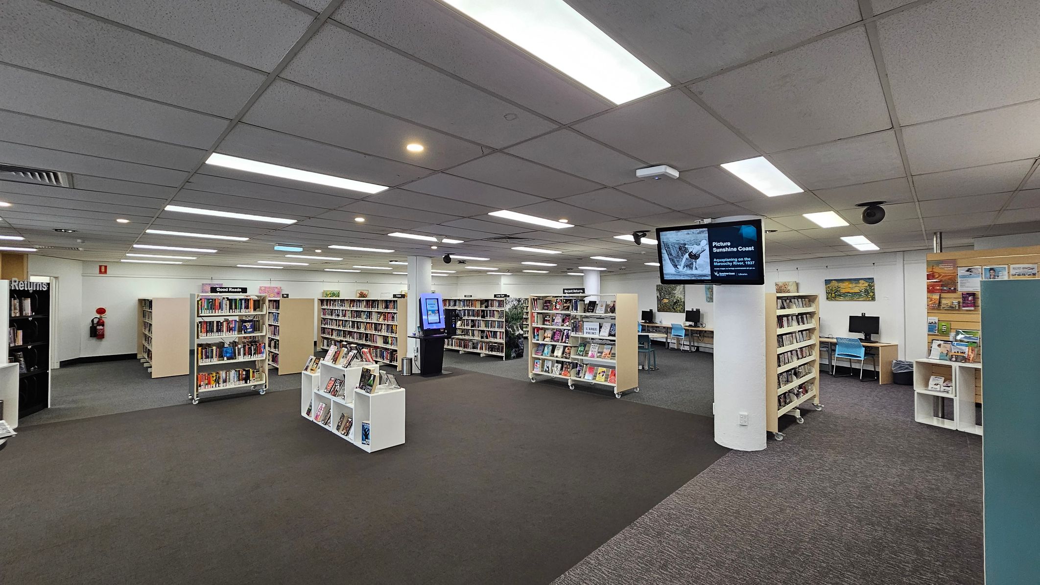 A library interior with grey carpeted flooring and a grid-patterned ceiling with fluorescent lights. In the centre, there are several bookshelves filled with books. On the right, a digital screen is mounted on a pillar, and computer stations with blue chairs are visible. On the left, a book return area is marked. The walls have posters and artwork, and a fire extinguisher is mounted near the corner.