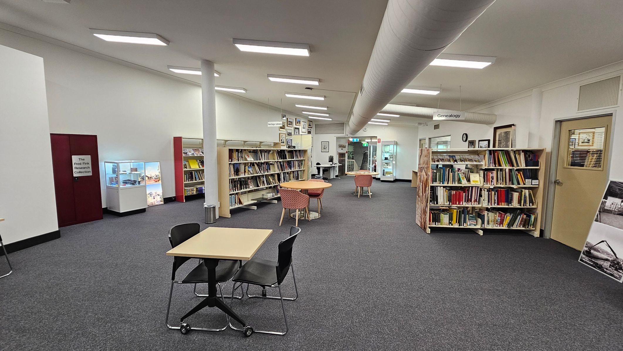 A library interior with grey carpeted flooring. On the left, there is a display case and a section labelled "The Fred Fink Research Collection." In the centre, there are bookshelves filled with books and a few round tables with red-patterned chairs. On the right, more bookshelves are visible, along with a door. The ceiling has fluorescent lights and a large ventilation duct running across. In the background, there are more tables and a computer station.