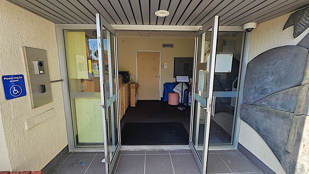 Glass double doors are open, leading into a building. On the left, there's a wall-mounted intercom system with a blue sign indicating wheelchair access. Inside, a beige door is visible at the back, with a small table and some items to the left. On the right, a decorative wall feature is partially visible. The ceiling is lined with grey panels, and a security camera is mounted near the entrance. The floor is tiled in dark grey.