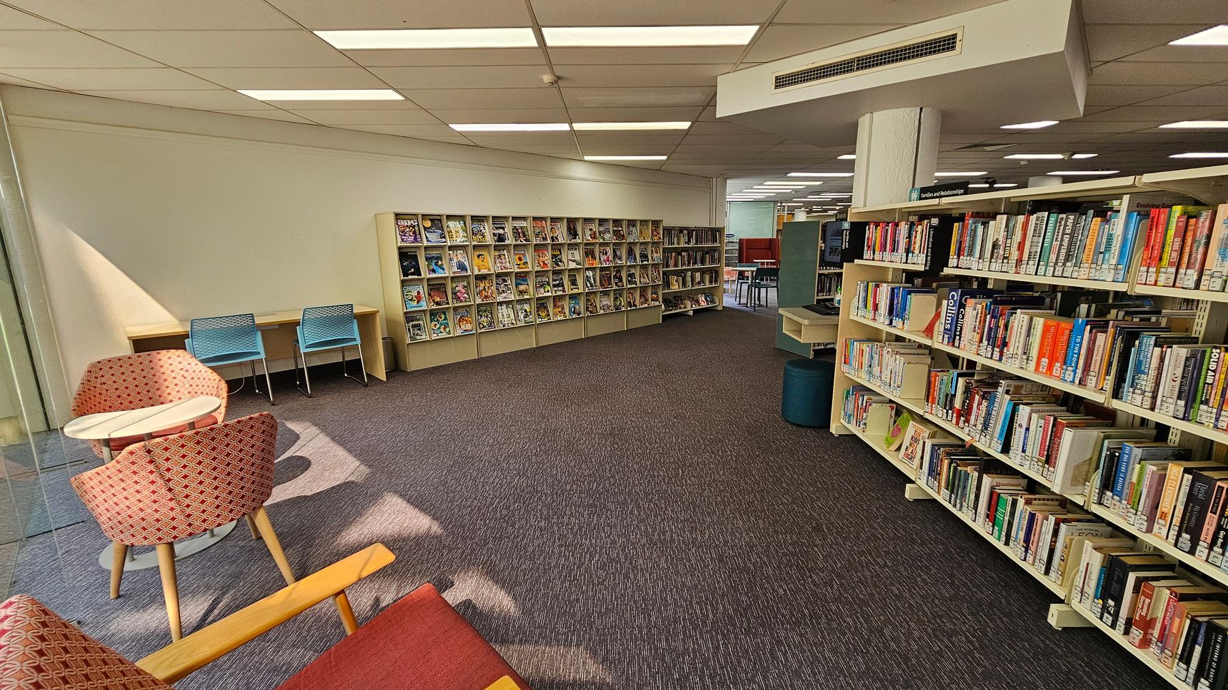 A library interior with bookshelves on the right filled with various books. In the middle, there are more shelves displaying magazines or comics. On the left, there are two blue chairs at a wooden desk and two red patterned chairs around a small round table. The floor is carpeted in a dark colour, and the ceiling has white panels with fluorescent lights.