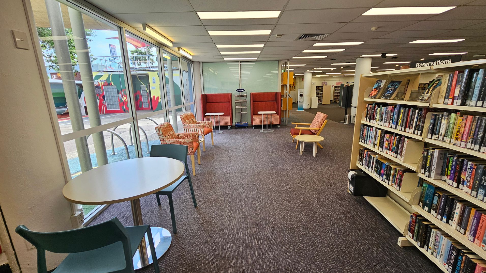 A library interior with a carpeted floor and a ceiling with fluorescent lights. On the left, large windows reveal an outdoor mural. In the centre, there are orange and patterned chairs with small round tables. On the right, a bookshelf labelled "Reservations" is filled with books. A small round table with a chair is near the window. The space is open and well-lit.