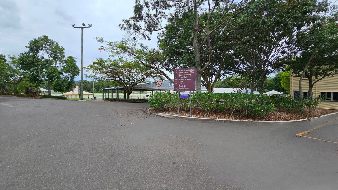 A two-storey building labelled "Main Pavilion" is in the centre, with a staircase leading to the entrance. The building is light-coloured with a gabled roof. On the left, there are trees and shrubs lining a path. On the right, more dense trees and several green bins are visible. The foreground features a large, empty paved area with some scattered leaves and a marked parking space.