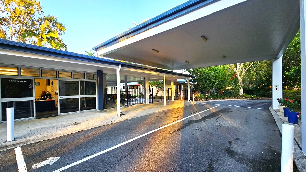 On the left, there is a building with large windows and a sign reading "Administration." Inside, there is a desk with a computer and some office supplies. On the right, there is a covered driveway with white support columns and a marked asphalt road. In the middle, the driveway leads to a garden area with trees and shrubs. The floor is a mix of concrete pavement near the building and asphalt on the driveway.