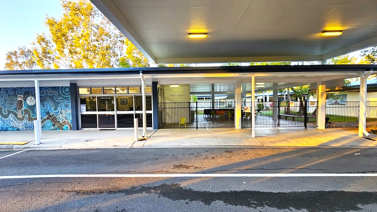 On the left, there is a building with a mural featuring blue and white patterns, and a glass entrance with double doors labelled "Administration." On the right, there is a covered area with white support columns and a metal fence enclosing a seating area with tables and chairs. In the middle, the covered area extends towards the background, revealing more of the building and trees. The floor is a mix of concrete pavement and asphalt.