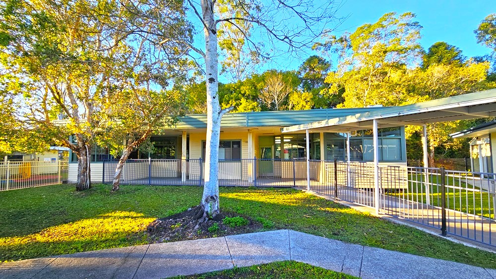 On the left, there is a covered walkway with a metal fence beside it. In the middle, a concrete path leads to a building with large windows and a slanted roof. On the right, another covered walkway with white pillars is visible. The background features tall trees with green foliage. The foreground has a grassy area with a concrete path intersecting it. The floor is a mix of grass and concrete paths, while the ceiling of the walkways is made of corrugated metal.