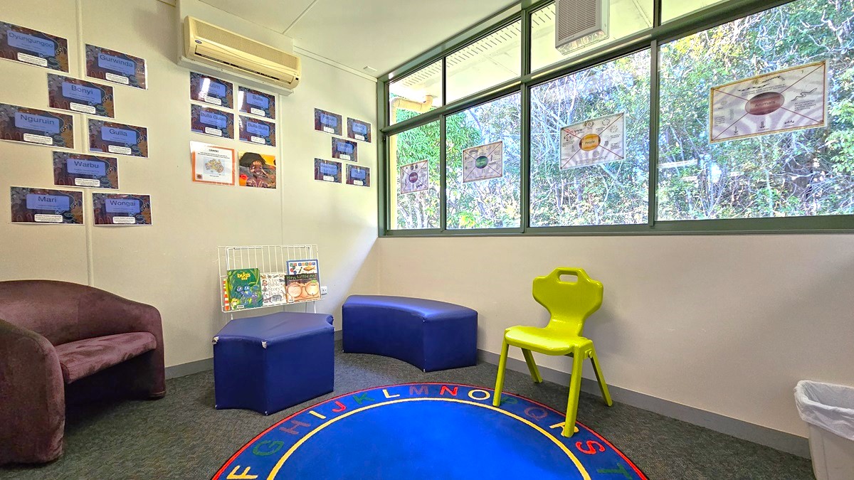 On the left, there is a brown armchair and a display of educational posters on the wall. In the middle, there are two blue cushioned seats and a small bookshelf with children's books. On the right, there is a green plastic chair and large windows with green frames, showing a view of trees outside. The background includes more educational posters and a white wall. The floor is carpeted in grey with a blue circular rug featuring the alphabet. The ceiling is white with an air conditioning unit.