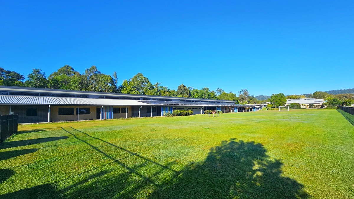 On the left, there is a black metal fence casting shadows on a large grassy field. In the middle, there is an expansive lawn with a few scattered trees casting shadows. On the right, there are several buildings with blue doors and a covered walkway supported by white pillars. The floor of the walkway is concrete, and the ceiling is metal. The background includes more trees and additional buildings.