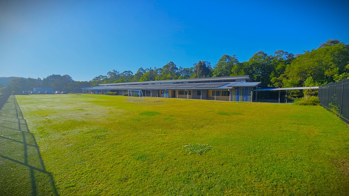 On the left, there is a black metal fence running along the edge of a large grassy field with a football goal. In the middle, there is an expansive lawn. On the right, there is a long building with a grey roof, blue doors, and a covered walkway supported by white pillars. The floor of the walkway is concrete, and the ceiling is metal. The background includes trees and additional buildings.