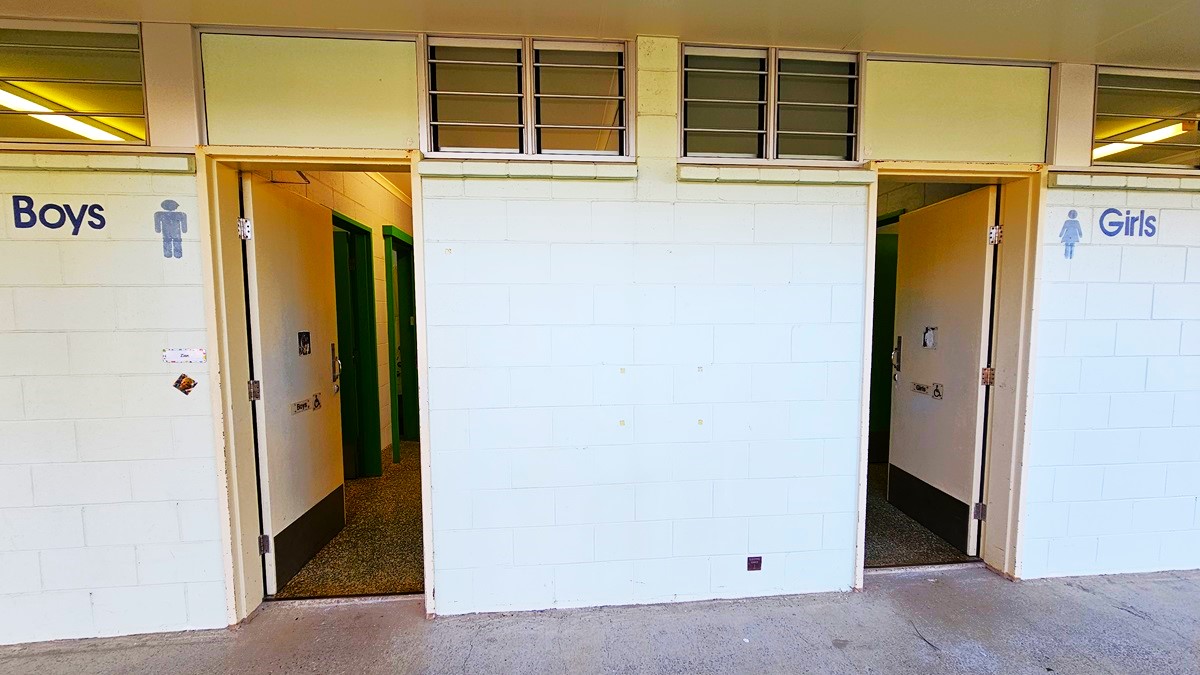 On the left, there is a doorway labelled "Boys" with a sign showing a male figure. On the right, there is a doorway labelled "Girls" with a sign showing a female figure. In the middle, there is a white brick wall with three small windows above it. The floor is concrete, and the ceiling is white with fluorescent lights visible through the doorways.
