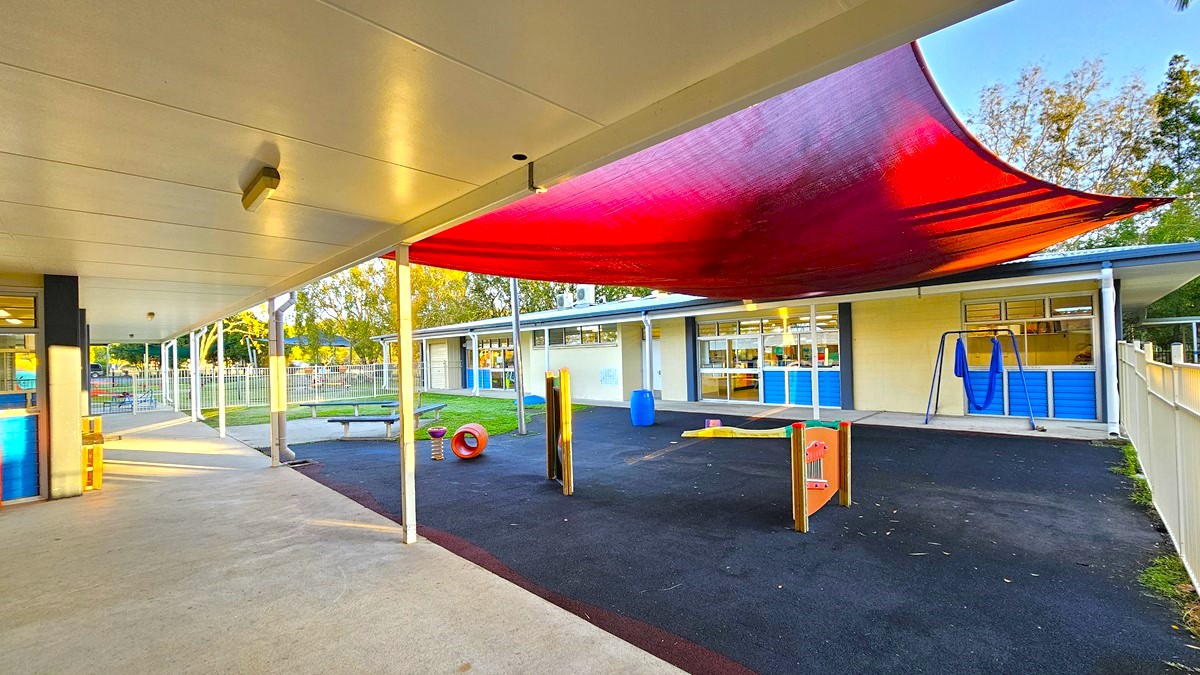 On the left, there is a covered walkway with white support columns and a metal fence enclosing a grassy area with a small garden. On the right, there is another covered walkway with white columns, leading to a building with large glass windows. In the middle, there is a concrete path connecting the two walkways, with grass and trees in the background. The floor is concrete, and the ceiling of the covered walkways is white with exposed beams.