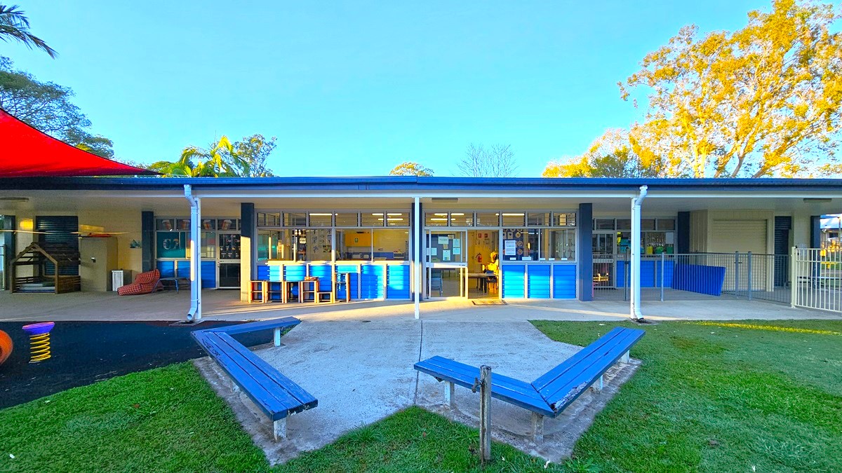 On the left, there is a covered area with white columns, a red shade sail, and playground equipment on a black rubber floor. On the right, there is a fenced area with blue railings and a concrete floor. In the middle, there is a building with large windows and blue doors, featuring a row of blue cabinets and shelves inside. The foreground has a concrete path flanked by blue benches on grass.