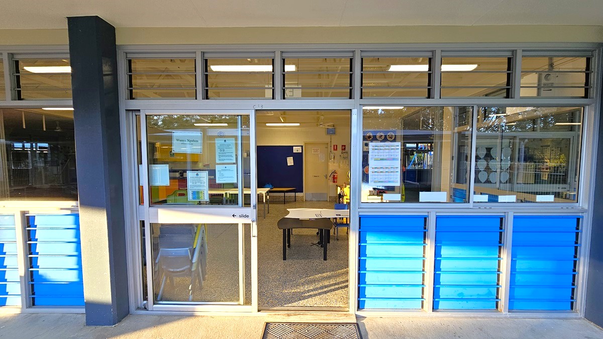 On the left, there is a black column next to a glass door with notices and a sign reading "Office." On the right, there are large glass windows with blue panels below them. In the middle, through the glass door, there is an office with a desk, chairs, and various notices on the walls. The floor inside is covered in grey carpet tiles, and the ceiling is white with exposed beams and fluorescent lighting. The exterior floor is concrete.