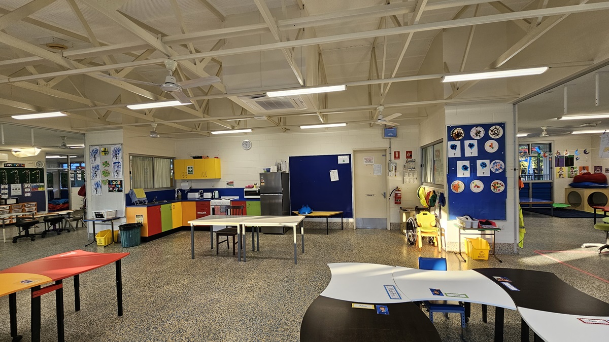 On the left, there are tables with colourful tops, a yellow and red cabinet, and a wall with children's artwork. On the right, there are more tables, a blue wall with circular art pieces, and a yellow walker. In the middle, there is a door with a clock above it, leading to a room with a refrigerator and storage cabinets. The floor is speckled grey, and the ceiling is white with exposed beams, fluorescent lights, and ceiling fans.