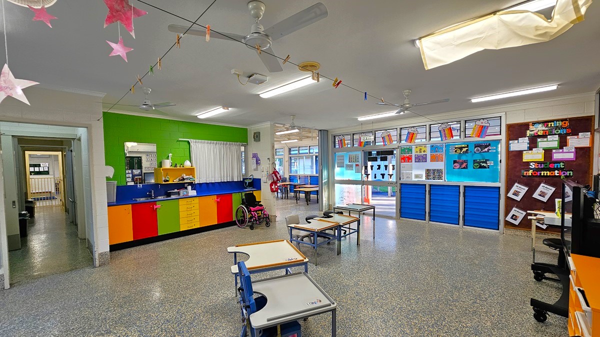On the left, there is a green wall with a window and a counter below it, featuring colourful cabinets and a pink wheelchair. On the right, there is a blue wall with bulletin boards displaying various papers and a "Student Information" sign. In the middle, there are tables and chairs, with a doorway leading to another room. The floor is speckled grey, and the ceiling is white with fluorescent lights, ceiling fans, and hanging decorations.
